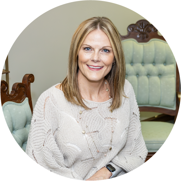 Headshot of Ashley Kopp, LCSW, a white middle aged woman wearing professional looking attire that is casual. In the background an antique chair gives a feeling of comfort and familiarity to her office.