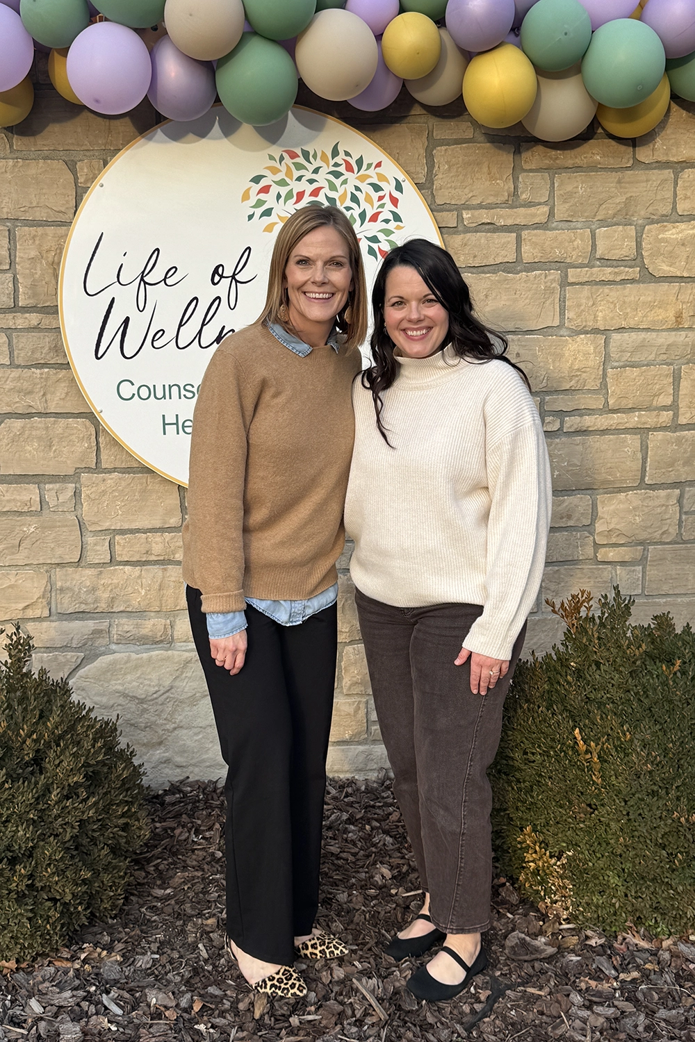 Photo of the team at Life of Wellness Counseling standing in front of their sign at their new location. Balloons decorate the stone wall behind them to celebrate their grand opening in January of 2026. Ashley Kopp is on the left with Miranda Fredrick on the right. Both middle aged women are smiling at the camera and are tucked together, showing a close working relationship as counselors in the practice located in Washington, Missouri.