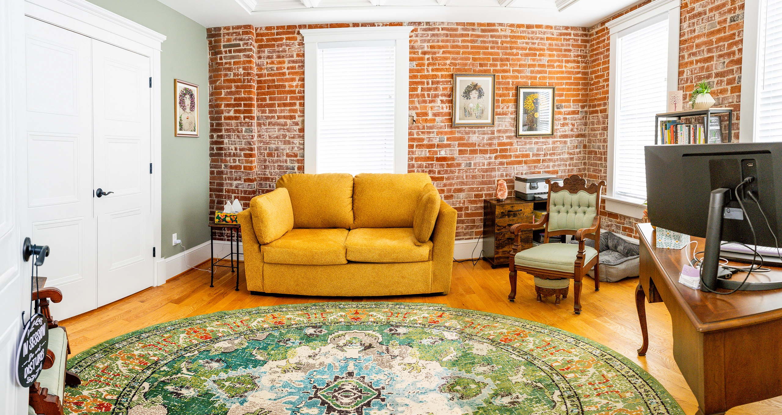 Image of the Life of Wellness Counseling office which is casual with a welcoming and professional vibe. A soft love seat couch provides a comfortable seating option while a cushioned wood chairs gives an alternative option. Desk space is on the right side of the room while a "In Session Do Not Disturb" sign hangs on the open door.
